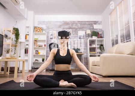 Caucasian young woman doing yoga in living room and wearing virtual reality goggles. Stock Photo