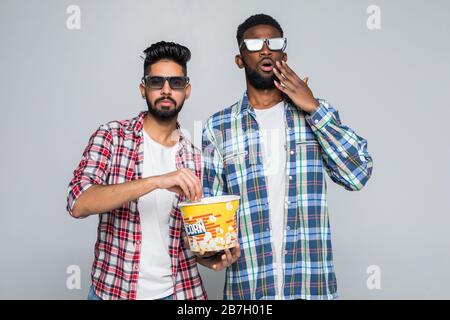Two mixed race men wear 3d glasses eat popcorn on gray background Stock Photo