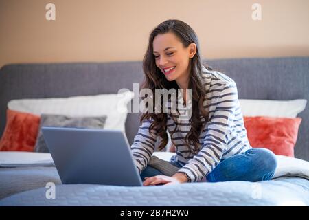 Happy young woman sitting on bed in her home and using laptop. Stock Photo