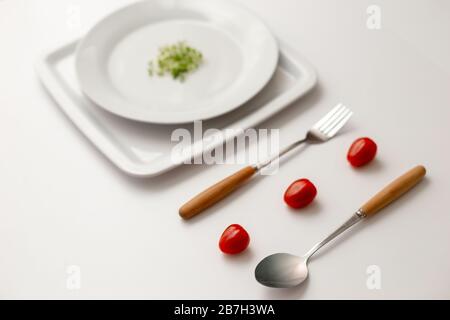 Three red cherry tomatoes between a fork and a spoon, small bunch of green watercress on a round white plates . Stock Photo