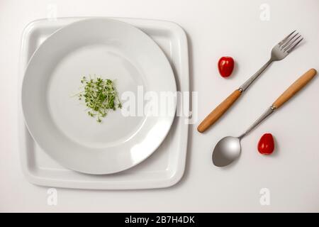 Two red cherry tomatoes between a fork and a spoon, small bunch of green watercress on a round white plates . Stock Photo
