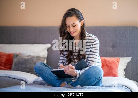Young woman is sitting on bed at home and writing diary. Stock Photo