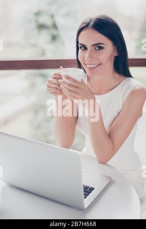 Cute young brunette is having tea at the cafe. She is happy, smiling. Her white outfit is very smart and stylish Stock Photo