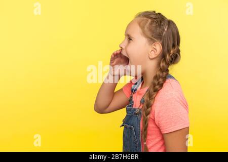 Attention! Side view of cute little girl with braid in denim overalls keeping hand near open mouth and screaming loudly, shouting announcement. indoor Stock Photo