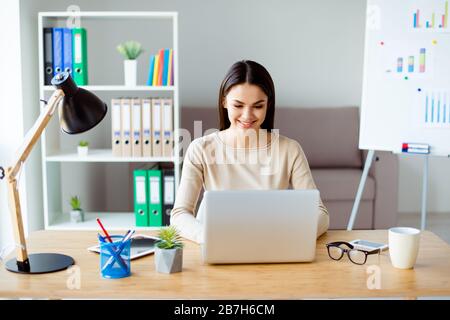 Cute beautiful young businesswoman sits at a desk in the office, types on a laptop and smiles Stock Photo