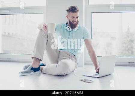 Happy cheerful smiling young man sitting on the floor drinking coffee and choosing music for listening Stock Photo