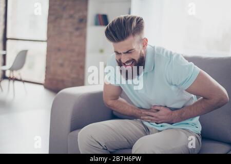 Sad young bearded man is suffering from stomach pain, ache is very hard. He is alone at home, sitting on couch Stock Photo