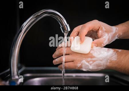 Wash your hands. a young guy washes his hands with soap under the faucet over a metal sink on a black dark background. Virus prevention. Hygienic proc Stock Photo