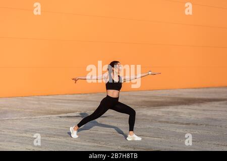 Fit young woman in tight sportswear, black pants and top, practicing yoga, standing in Virabhadrasana ii warrior pose and meditating, doing stretching Stock Photo