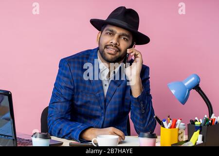 Joyful stylish indian entrepreneur wearing formal wear, black hat, talking on mobile phone, smiling broadly, looking to side, sitting at desk in offic Stock Photo