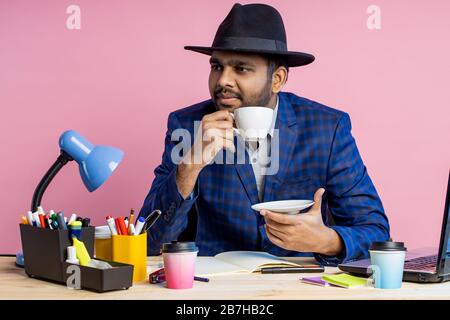 Close up cropped shot of young unshaven indian businessman in formal blue checkered suit, black hat , sitting at workplace, thinking about something, Stock Photo
