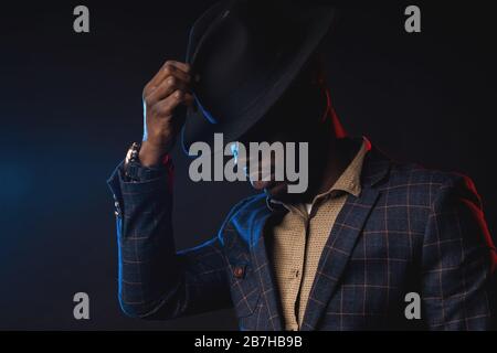 Side view of cool dark skinned man in stylish suit, touching his black hat, looking down. Closeup portrait of fashionable african american male model Stock Photo