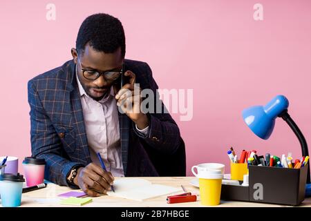 Serious afro american businessman in formal wear, glasses, talking on mobile phone, looking at notepad, holding pen, sitting at desk in office. Job, b Stock Photo