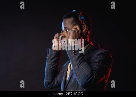 Closeup shot of african american young unshaven man listening music in headphones, looking down, posing in dark. Portrait of stylish DJ with headset, Stock Photo