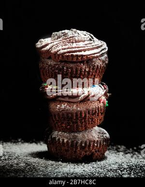 A pile of chocolate cupcakes decorated with powdered sugar on black background. Vertical portrait of the muffins. Perfect sweet food for celebrations. Stock Photo