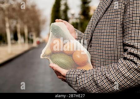 closeup of a man in the street carrying a textile reusable mesh bag, used to buy groceries in bulk, full of fruit and vegetables, as a measure to redu Stock Photo