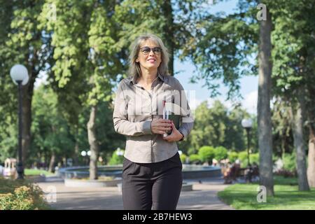 Business confident mature woman walking with documents outdoor. Beautiful blonde female in glasses, gray shirt, sky park background Stock Photo