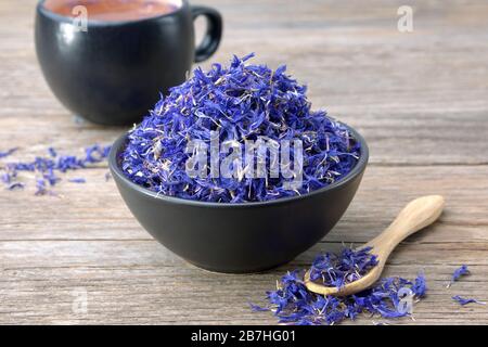 Black ceramic bowl of dry blue cornflowers petals and herbal tea cup on wooden table. Herbal medicine. Stock Photo