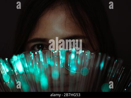 Diverse Asian girl looking through futuristic, teal coloured bokeh - Millennial woman looking at camera with advanced fibre optic technology - Next generation super fast internet and optical tech concept Stock Photo