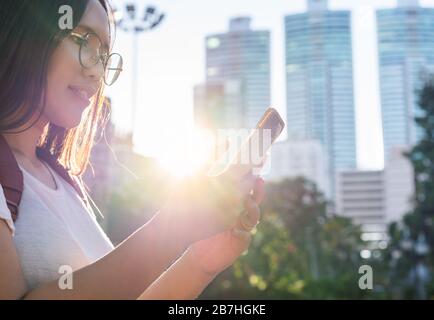 Diverse Asian girl looking at smart phone, in urban city park with bright afternoon sunshine - Young millennial hipster student woman holding mobile device outdoors - One person solo travel concept Stock Photo