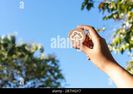 Diverse Asian female hand, holding up wood disc with cloud computing upload icon - Cloud computer file transfer symbol with blue sky - Remote connectivity, data storage and digital security concepts Stock Photo