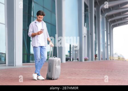 Online Taxi App Concept. Young Man Standing Near Airport Using Smartphone Stock Photo