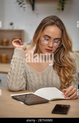 Girl blogger in glasses, beige sweater. Sitting in kitchen at table with smartphone and tablet on it. Going to write something in notebook. Close-up Stock Photo