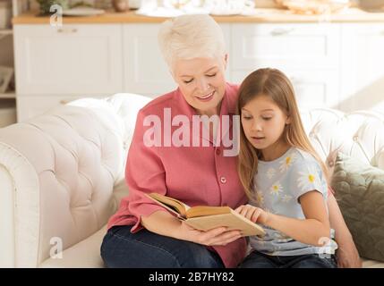 Elderly lady and her granddaughter with interesting book on sofa at home Stock Photo
