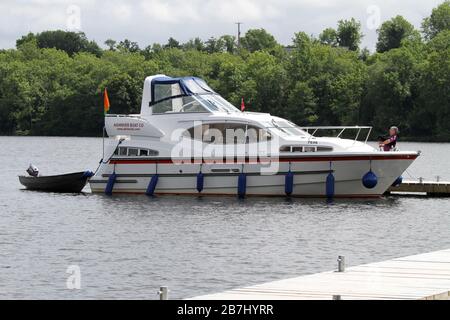 A man playing bagpipes on a wooden jetty beside a white Aghinver cruiser on Lower Lough Erne, County Fermanagh on a bright summer day. Stock Photo