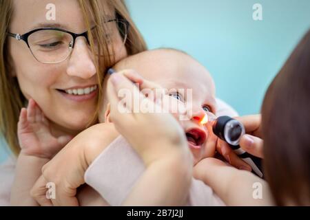 Doctor pediatrist examining childs nose with otoscope. Mom holding baby with hands while health checkup. Medicine, healthcare, pediatry and people Stock Photo