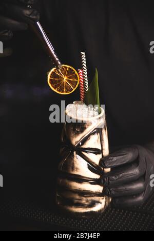 Tiki Drink Cocktail on the bar. Bartender with gloves decorates the drink with dried orange. Stock Photo