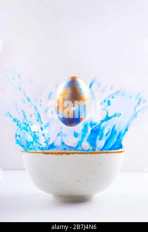 A painted pearl Easter egg in blue and gold flies in a bowl of blue paint. Greeting card for the holiday. Stock Photo