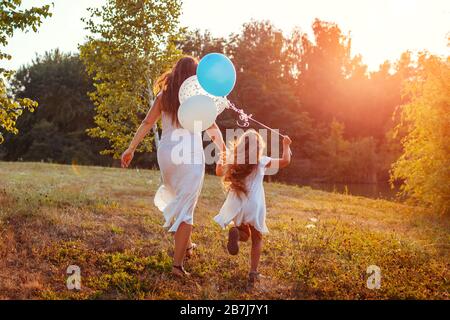 Mother's day. Little girl running with mother and holding baloons in hand. Family having fun in summer park