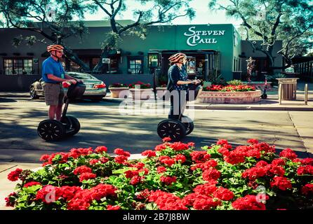 Foreground of red begonias with Caucasian woman and man wearing orange helmets riding Segway personal transporters in old town Scottsdale, AZ, USA Stock Photo