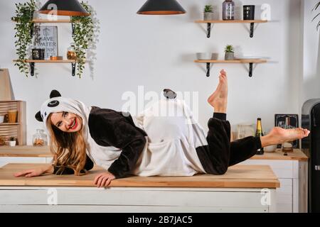 Lady dressed up in plush pajama in form of cartoon character panda is having fun at bachelorette party, posing on kitchen table, smiling. Close-up. Stock Photo