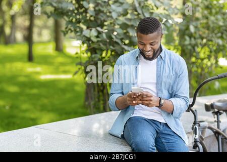 Black Hipster Guy Using Smartphone Outdoors, Sitting On Bench In Park Stock Photo