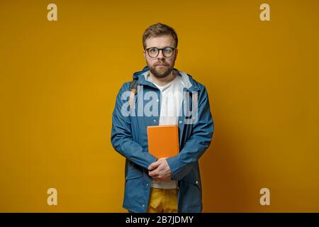 Attractive student or young scientist is standing with a book in hands, in a raincoat and calmly looking at the camera. Stock Photo