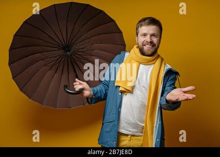 Joyful man in a bright raincoat and knitted scarf stands with an open umbrella, checks the rain with hand, on a yellow background. Stock Photo