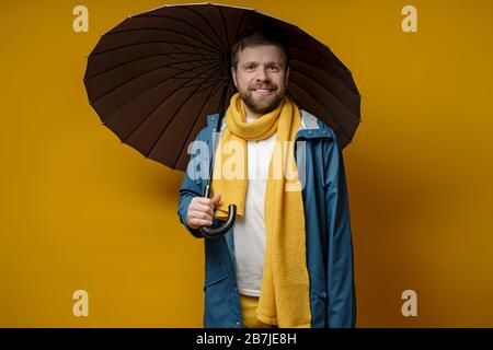 Smiling man in a bright raincoat and knitted scarf stands with an open umbrella, on a yellow background. Stock Photo