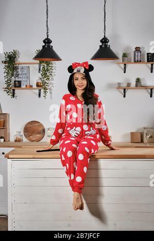 Girl dressed up in red plush pajama in form of cartoon character mouse is having fun at hen-party. She is smiling, sitting on a wooden kitchen table. Stock Photo