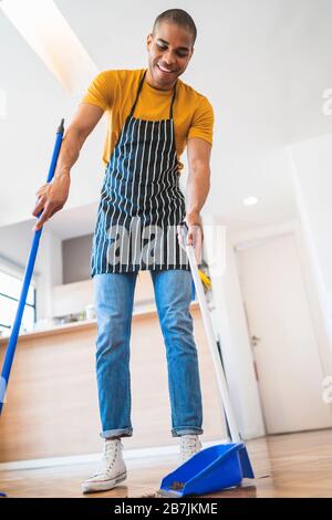 Portrait of young latin man sweeping wooden floor with broom at home. Cleaning, housework and housekeeping concept. Stock Photo