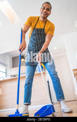 Portrait of young latin man sweeping wooden floor with broom at home. Cleaning, housework and housekeeping concept. Stock Photo