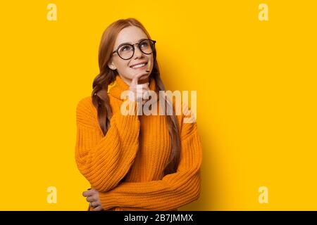 Thoughtful ginger caucasian lady is touching her chin while looking through eyeglasses and posing on a yellow background in a nice sweater Stock Photo