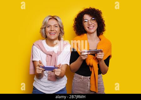 Curly haired caucasian woman with eyeglasses is posing in her blonde sister while playing some mobile games on a yellow background Stock Photo