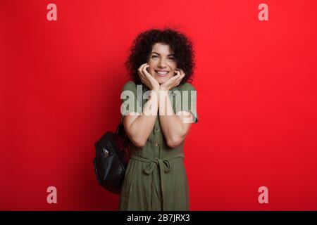 Curly haired caucasian student is cheering on a red background while holding a bag and touching her chin with both hands Stock Photo