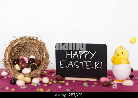Bird nest full of easter almond eggs, decorative chick and blackboard with the text Happy Easter Stock Photo