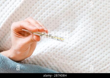 Sick man holding a oral mercury thermometer in his hand, showing high temperature, lying in bed, copy space. Common cold, illness, flu season concept Stock Photo