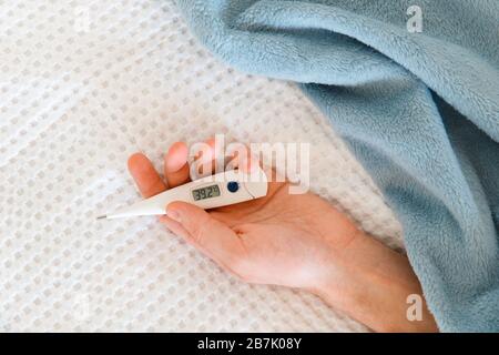 Closeup of man hand holding a digital thermometer, lying on the bed, indoors. High body temperature, cold, flu epidemic concept Stock Photo