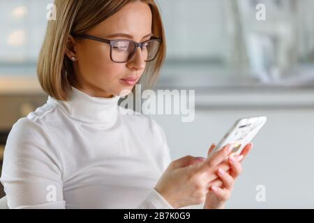 Serious businesswoman wears white turtleneck sweater and glasses using modern smartphone in marble case, browse internet, buyng goods online, blurred Stock Photo