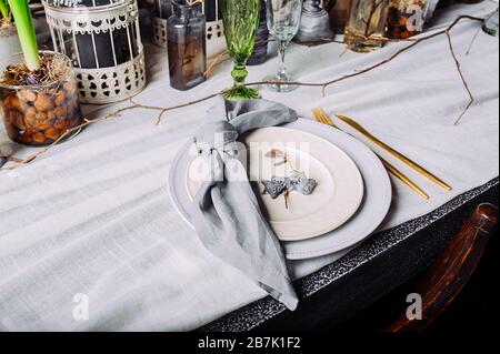 Table setting in a mysterious spring style with live plants and branches. White plates with gold Cutlery and a blue cotton napkin tied in a knot. Stock Photo